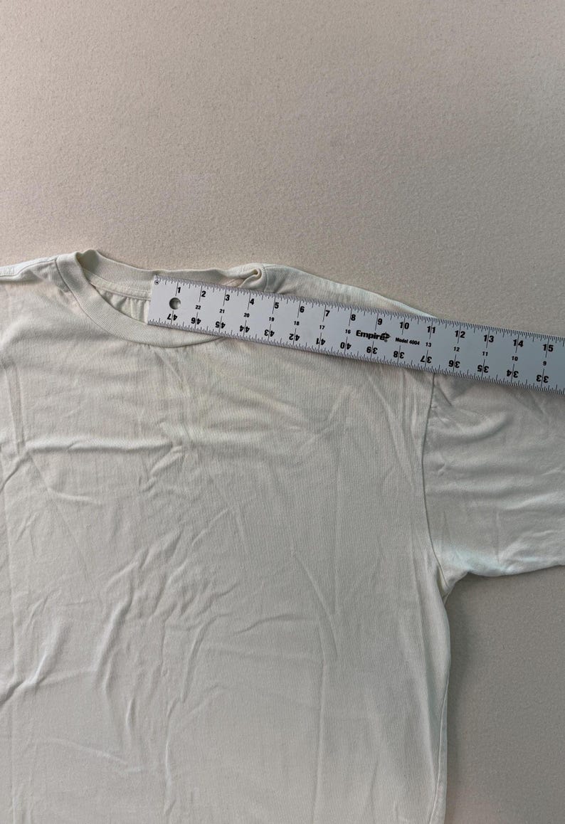 May include: A cream-colored t-shirt with a crew neck. A ruler is placed across the shoulder of the shirt, showing measurements in centimeters. The shirt is made of a soft, plain fabric.