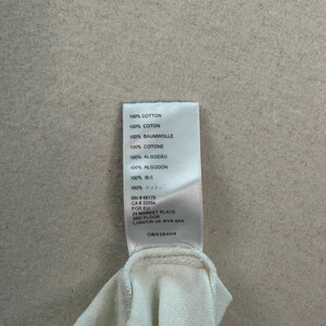 May include: A close-up of a white fabric garment with a tag attached. The tag lists the fabric composition as 100% cotton in multiple languages. The tag also includes a RN and CA number, and the address of the manufacturer.