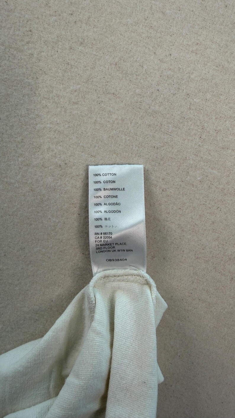 May include: A close-up of a white fabric garment with a tag attached. The tag lists the fabric composition as 100% cotton in multiple languages. The tag also includes a RN and CA number, and the address of the manufacturer.