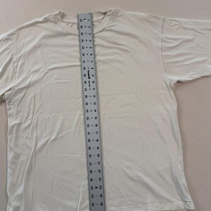 May include: A cream-coloured t-shirt with a crew neck is laid flat. A ruler is placed vertically on the shirt, measuring its length. The shirt appears to be a size large, with a length of approximately 61 cm.