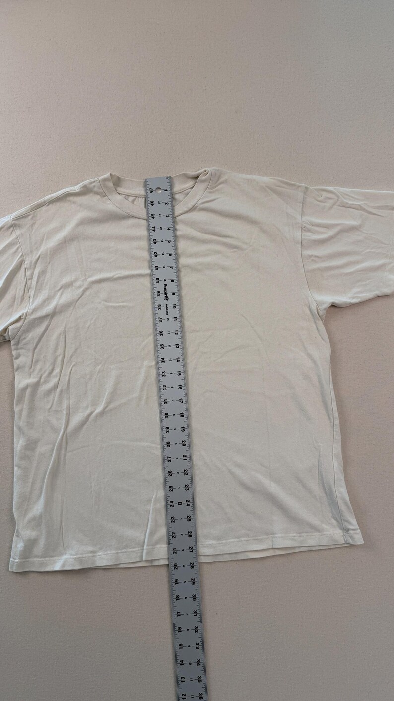 May include: A cream-coloured t-shirt with a crew neck is laid flat. A ruler is placed vertically on the shirt, measuring its length. The shirt appears to be a size large, with a length of approximately 61 cm.