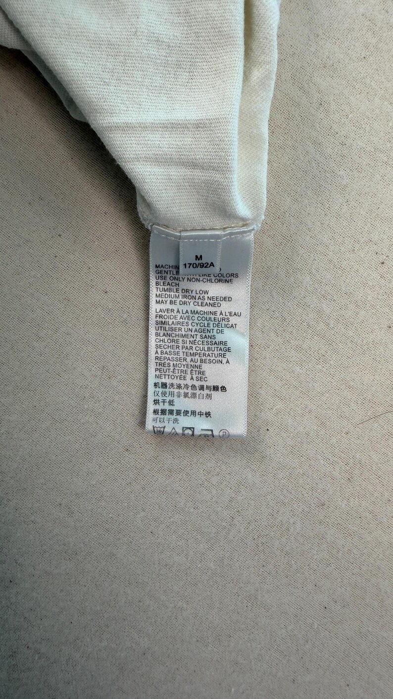 May include: A close-up of a white fabric with a tag attached. The tag has text in multiple languages, including English and French, with washing instructions and a size indicator.