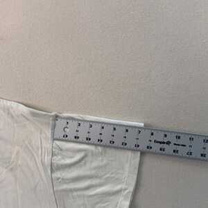 May include: A white t-shirt sleeve is measured with a ruler. The ruler shows measurements in inches. The sleeve is approximately 15 cm long.