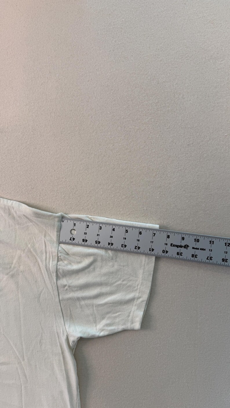 May include: A white t-shirt sleeve is measured with a ruler. The ruler shows measurements in inches. The sleeve is approximately 15 cm long.