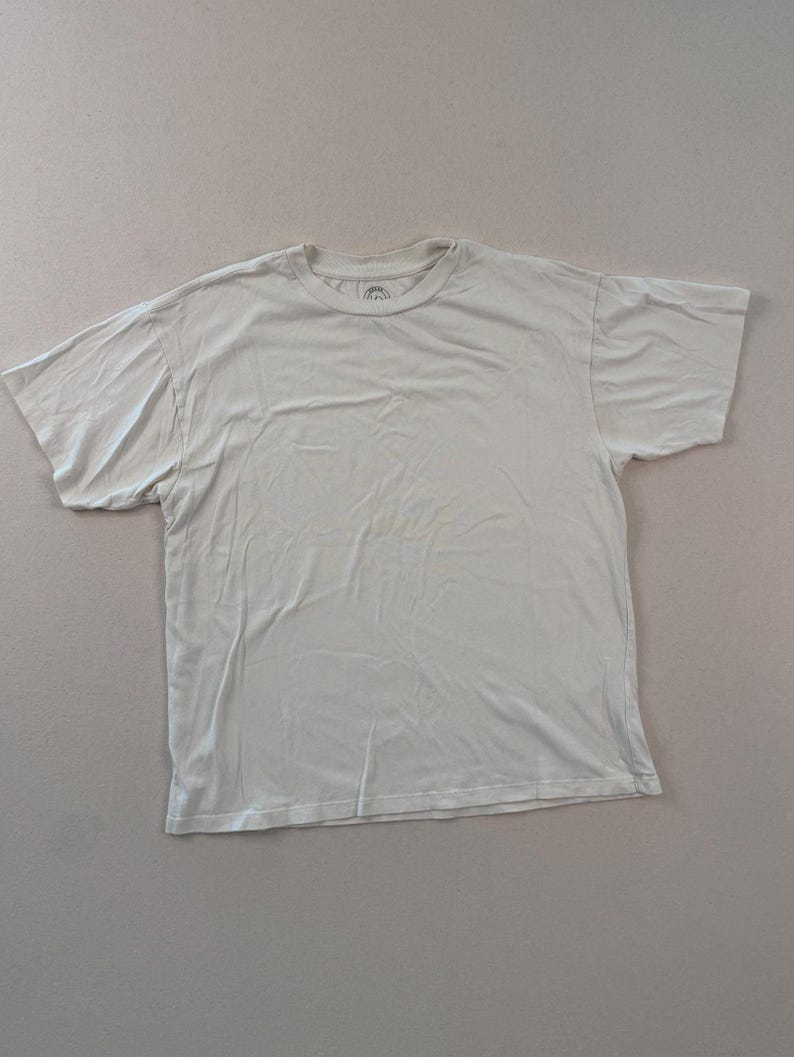 May include: A pale-coloured, short-sleeved t-shirt. The shirt has a crew neck and appears to be made of a soft material. The shirt is laid flat on a neutral surface.