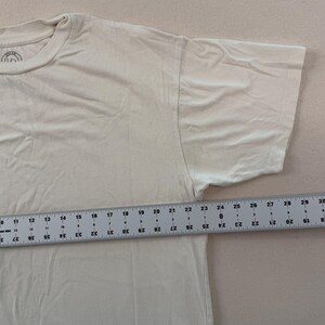 May include: Cream-coloured short-sleeved t-shirt. A ruler shows the shirt's length is approximately 76 cm. The shirt has a crew neck and a simple design, suitable for casual wear. The fabric appears to be soft and comfortable.