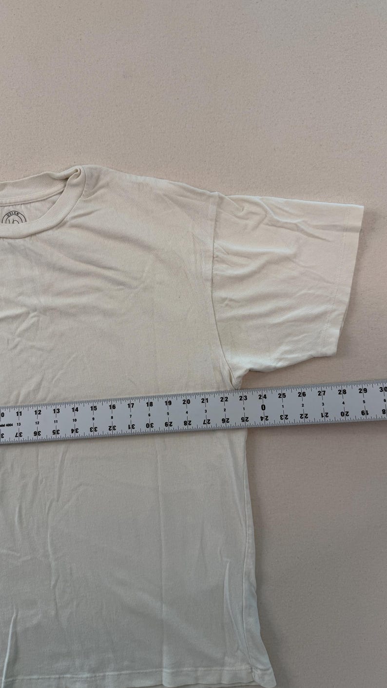 May include: Cream-coloured short-sleeved t-shirt. A ruler shows the shirt's length is approximately 76 cm. The shirt has a crew neck and a simple design, suitable for casual wear. The fabric appears to be soft and comfortable.