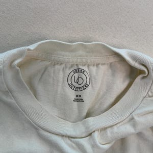 May include: Cream-coloured t-shirt with a crew neck. The shirt features a circular logo with the words "Urban Outfitters" and the size "M-M" printed on the inside collar. The tag also says "Made in India" and "Fabriqu&eacute; en Inde".