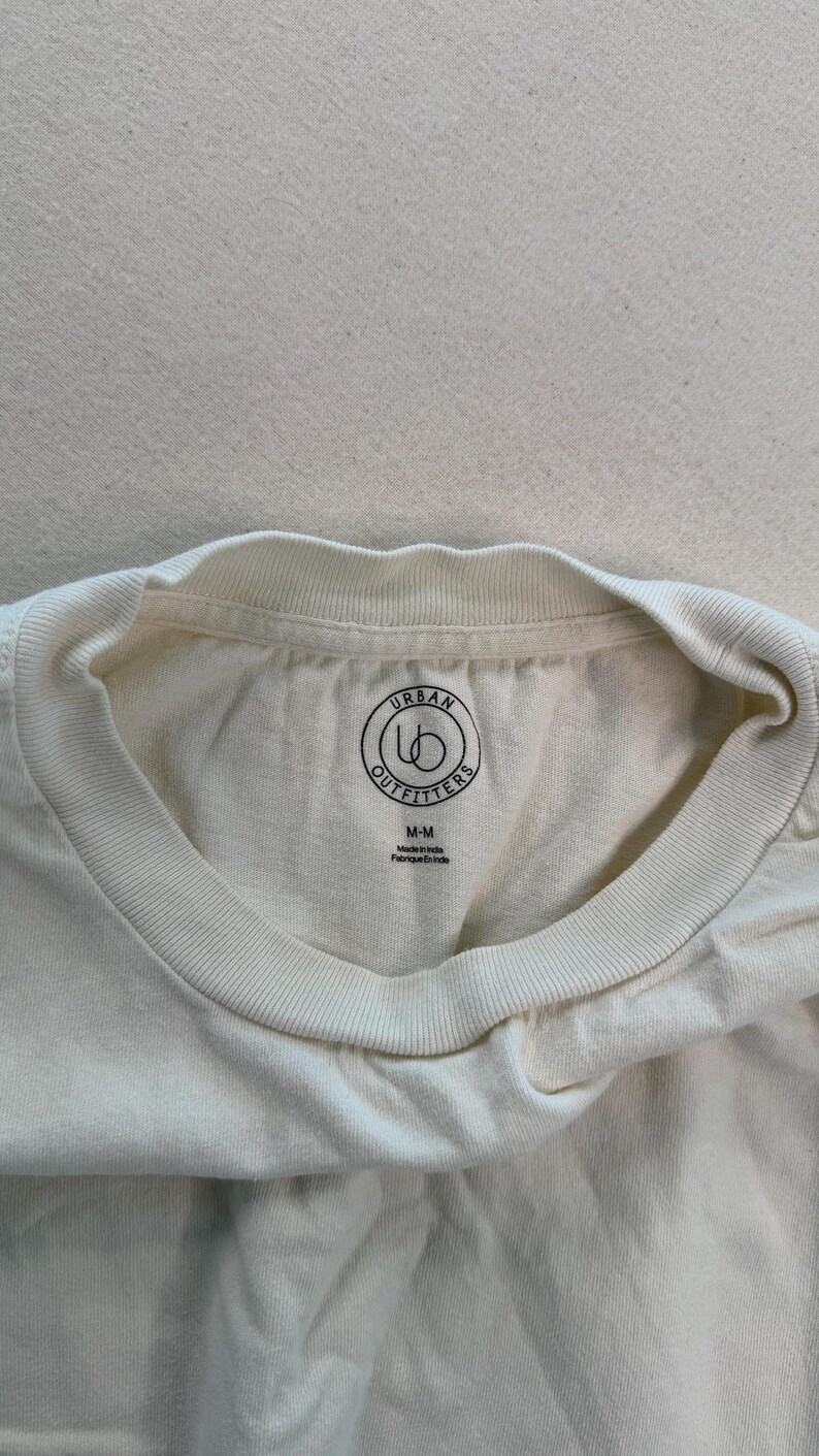 May include: Cream-coloured t-shirt with a crew neck. The shirt features a circular logo with the words "Urban Outfitters" and the size "M-M" printed on the inside collar. The tag also says "Made in India" and "Fabriqu&eacute; en Inde".