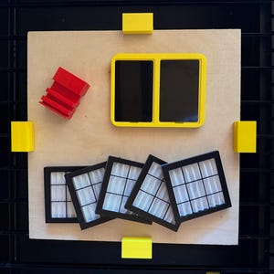 May include: A collection of geometric shapes in primary colors arranged on a light wood surface. Includes a yellow rectangular frame with two black rectangles, five black framed squares with white mesh, red stacked blocks, and yellow blocks.