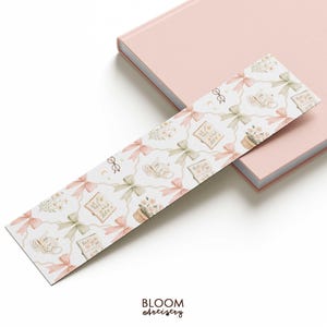 May include: A decorative bookmark with a repeating pattern of pink and green bows, teacups, books, and potted flowers on a white background. The bookmark is placed on a pink book. The text "BLOOM Artistry" is at the bottom.