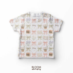 May include: White t-shirt with a repeating pattern of pink and green bows, books, eyeglasses, lavender, and potted plants. The shirt has a button closure at the neckline and the text "BLOOM artistry" at the bottom.