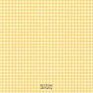 Può includere: Un motivo senza cuciture di quadri vichy gialli e bianchi. Il design presenta una griglia ripetuta di quadrati, creando un'estetica classica e allegra. Le parole "BLOOM artistry" sono stampate in un carattere di scrittura in basso a destra.