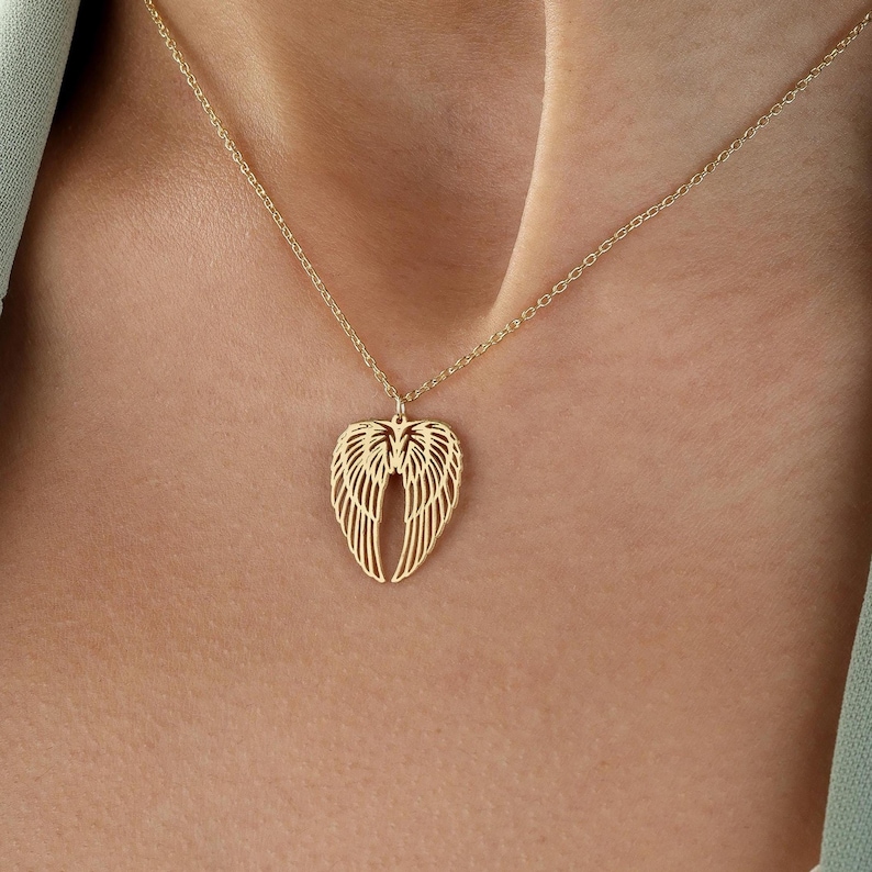 May include: A gold-colored necklace featuring a pendant shaped like angel wings. The wings are intricately detailed with delicate lines, suspended from a fine chain. The necklace is displayed against a skin tone background.