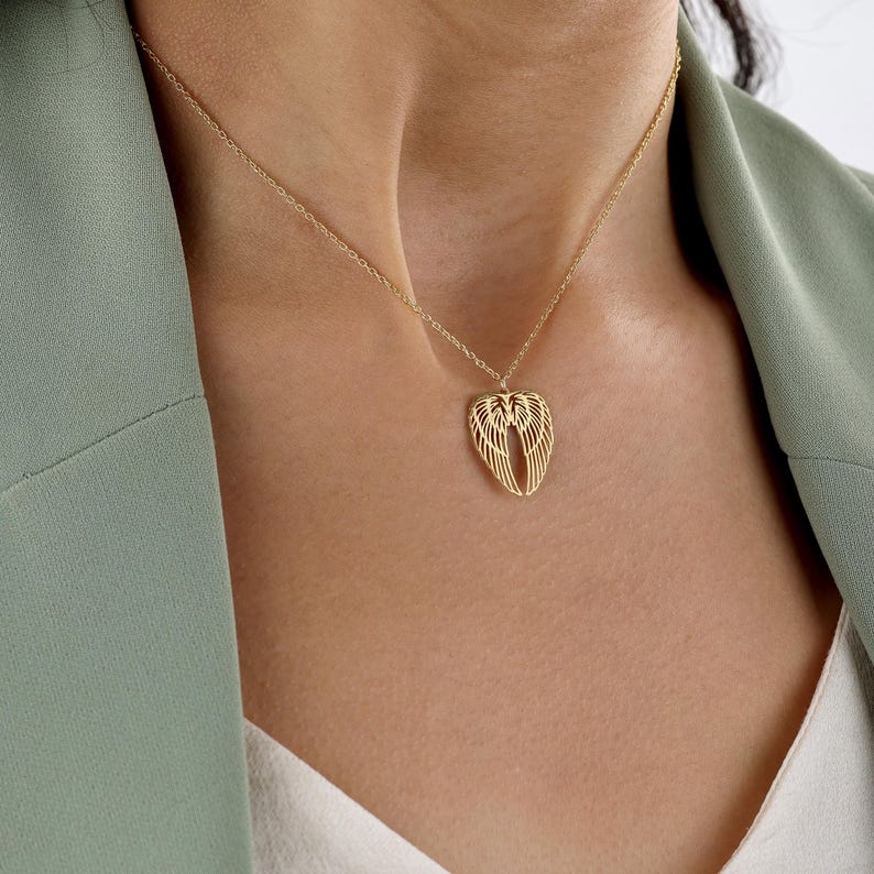 May include: A gold-colored necklace featuring a pendant shaped like angel wings. The wings are detailed with delicate lines, and the necklace is displayed on a person wearing a green blazer.