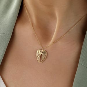 May include: A gold-colored necklace with a pendant shaped like angel wings. The wings are detailed with multiple lines, and the necklace is worn against a person's skin and a green blazer.