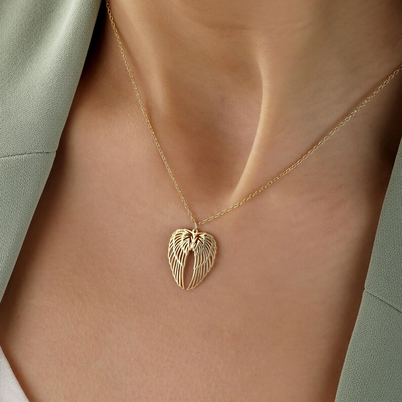 May include: A gold-colored necklace with a pendant shaped like angel wings. The wings are detailed with multiple lines, and the necklace is worn against a person's skin and a green blazer.