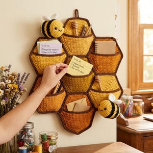 Puede incluir: Un organizador de pared con forma de panal hecho de hilo de ganchillo en tonos amarillo, naranja y marrón. Cuenta con múltiples bolsillos, dos abejas de peluche adjuntas y contiene notas con el texto To do do: Organize supplies Bee-utiful day!