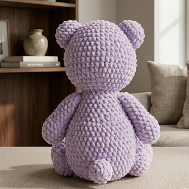 May include: A handmade, lavender-colored crochet teddy bear. The bear is seated, showcasing its round head, small ears, and detailed textured body. The soft, plush texture is visible, suggesting a cozy, comforting toy. The bear is a charming, handcrafted item.