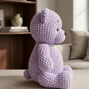 May include: A large, lavender-colored crocheted teddy bear with black eyes and nose. The plush toy is sitting on a neutral-colored surface. The bear has a textured, chunky knit appearance.