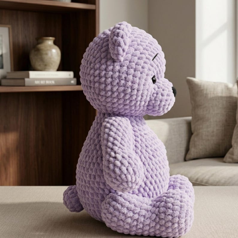 May include: A large, lavender-colored crocheted teddy bear with black eyes and nose. The plush toy is sitting on a neutral-colored surface. The bear has a textured, chunky knit appearance.