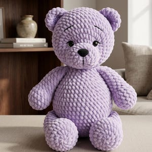 May include: A handmade, lavender crochet teddy bear with a black nose and eyes. The soft toy has a textured appearance and is sitting upright. The bear's arms and legs are rounded, and it has small, rounded ears. The bear is a soft toy.