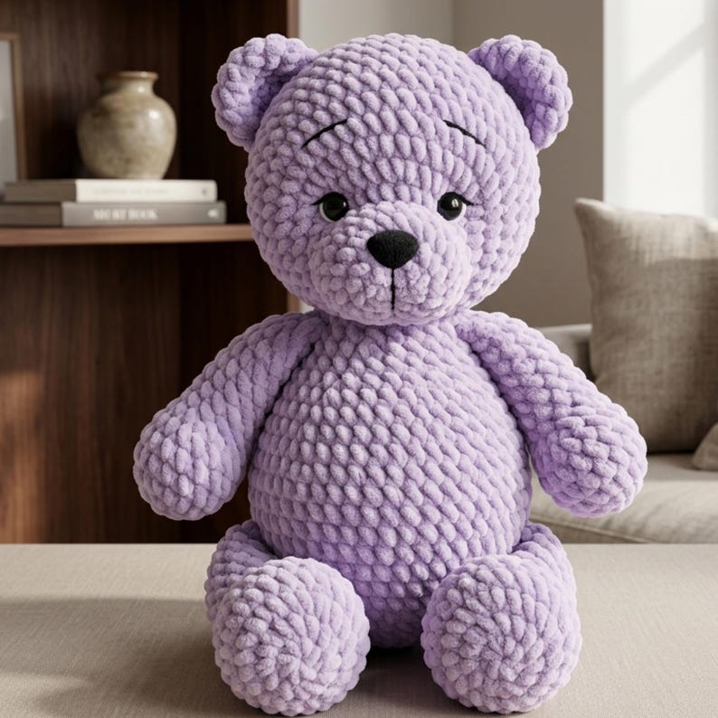 May include: A handmade, lavender-colored crochet teddy bear with a black nose and eyes. The plush toy has a textured, soft appearance and is sitting upright. The bear's arms and legs are rounded, and it has small, rounded ears.