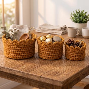 May include: Three round, mustard-yellow crocheted baskets with scalloped edges. The largest basket holds a light-colored cloth and a wooden scoop. The middle basket contains gold-wrapped candies and a stone. The smallest basket holds pinecones and a key.