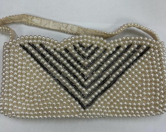May include: A rectangular handbag covered in white faux pearls with a scalloped top edge. The front features a V-shaped pattern created with black cylindrical beads. The bag includes a matching white pearl-covered strap attached to the sides.