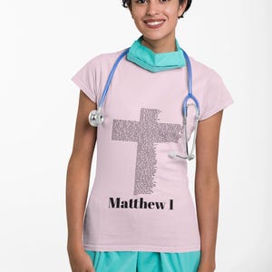 May include: A light pink t-shirt with a cross made of text and the words "Matthew 1" in black. The person is wearing a teal face mask and a blue stethoscope. They are also wearing teal scrubs.