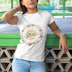Könnte beinhalten: Weißes T-Shirt mit einem Sonnendesign mit Gesicht, umgeben von einem floralen Kranz. Die Sonne hat ein gelbes Gesicht mit geschlossenen Augen und einem lächelnden Mund. Der Kranz enthält gelbe und rosa Blumen und grüne Blätter. Die Person trägt eine blaue Jeans.