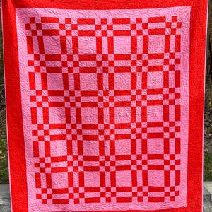 May include: A handmade quilt with a vibrant color scheme. The quilt features a pink background with a grid pattern in bright red. A solid red border frames the design. The quilt is likely made of cotton or a similar fabric.