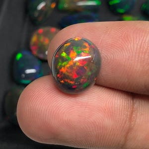 Può includere: Una gemma di opale nero ovale lucidata che presenta vivaci bagliori di colore rosso, arancione e verde. La pietra è liscia e arrotondata, mostrando una tonalità di base scura con intensi riflessi di luce interni sulla sua superficie.