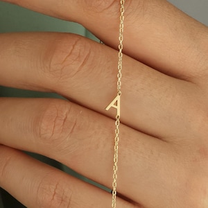 May include: A delicate gold necklace featuring the letter "A". The pendant hangs from a fine chain, creating a minimalist and elegant look. The necklace is shown against a soft green background.
