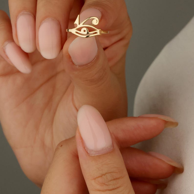May include: A gold-colored ring featuring an openwork design of the Eye of Horus. The ring is held up against a blurred background. The ring's design is a stylized eye with a curved line above it.