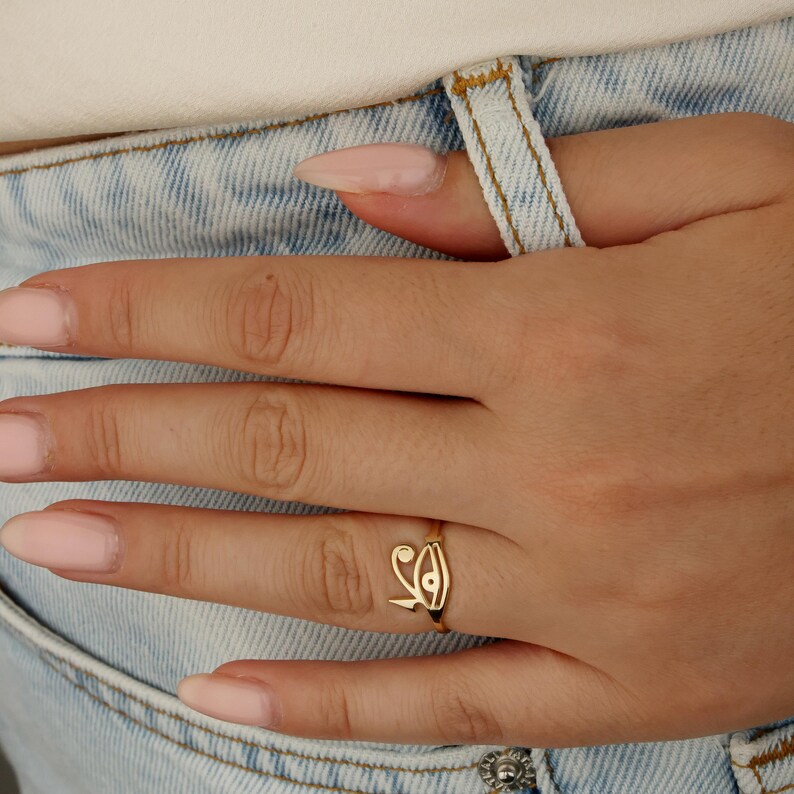 May include: A gold-colored ring featuring an eye-like design. The ring is worn on a finger, set against a backdrop of light blue denim jeans and a white top. The ring's design is intricate and detailed.