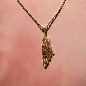 Può includere: Collana con pendente color oro con un design intricato e fluido. Il pendente pende da una delicata catena su uno sfondo rosa. Il design del pendente presenta motivi e forme a vortice, creando un aspetto elegante e artistico.