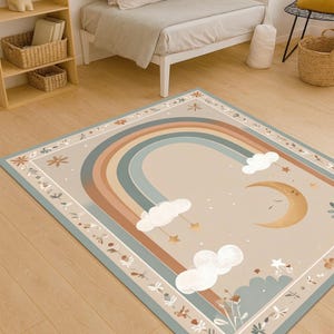 Puede incluir: Una alfombra rectangular con un dise&ntilde;o de arco&iacute;ris en colores apagados beige, marr&oacute;n y azul. La alfombra presenta una luna creciente, nubes y estrellas. El borde tiene un patr&oacute;n floral. Esta alfombra es adecuada para una habitaci&oacute;n infantil.