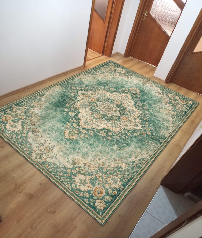 K&ouml;nnte beinhalten: Ein rechteckiger Teppich im Used-Look mit einem zentralen Medaillon-Design. Der Teppich hat eine dominierende tealgr&uuml;ne Farbe mit cremefarbenen und orangefarbenen Blumenmustern. Er liegt auf einem Holzboden und verleiht dem Raum W&auml;rme.