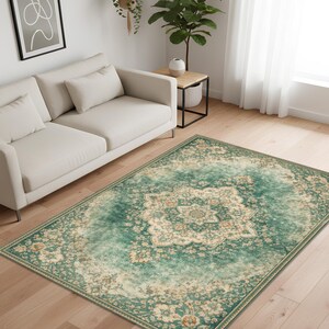K&ouml;nnte beinhalten: Ein rechteckiger Teppich mit einem Used-Look-Design im Vintage-Stil. Der Teppich hat ein zentrales Medaillonmuster in Creme und Beige, umgeben von einem t&uuml;rkis-gr&uuml;nen Rand mit floralen Motiven. Der Teppich liegt auf einem Holzboden.