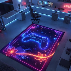 Puede incluir: Una alfombra rectangular con un borde rosa y azul neón. La alfombra presenta un diseño de mando de videojuego azul brillante en el centro, con un fondo estrellado. Dos flores estilizadas y una línea naranja brillante completan el diseño.