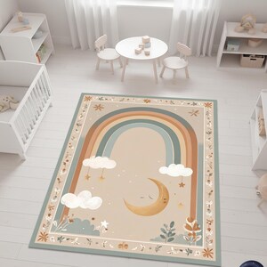 Puede incluir: Una alfombra rectangular con un dise&ntilde;o caprichoso que presenta un arco&iacute;ris, nubes, estrellas y una luna creciente. La alfombra tiene un fondo beige con un borde de patrones florales y de hojas. El arco&iacute;ris tiene tonos azules, marrones y beige.