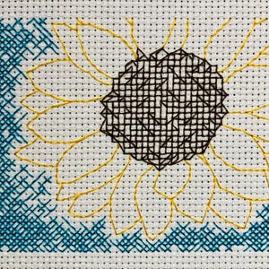 Puede incluir: Punto de cruz de un girasol con pétalos amarillos y un centro marrón oscuro. El fondo es blanco con un borde azul verdoso en la esquina superior izquierda. El girasol está centrado.
