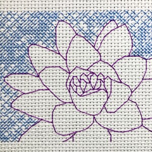 Puede incluir: Bordado a punto de cruz de una flor morada con pétalos detallados y un centro texturizado. La flor se encuentra sobre un fondo azul bordado a punto de cruz. El diseño está sobre una tela blanca punteada, mostrando los detalles cosidos a mano.