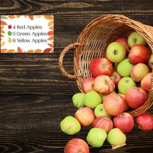 May include: A woven wicker basket tipped on its side, spilling a collection of red, green, and yellow apples. A rectangular sign with an autumn leaf border displays the text: 4 Red Apples, 5 Green Apples, 6 Yellow Apples.