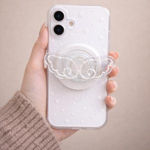 Op de afbeelding: Transparante telefoonhoes met een witte vlindervormige grip versierd met strass. De hoes heeft een sterrenpatroon en een witte cirkelvormige telefoongrip. De telefoon heeft twee cameralenzen.