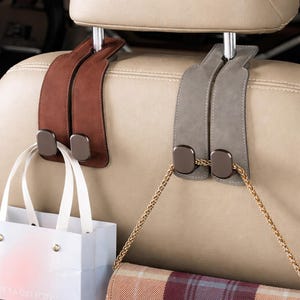 May include: Two car seat headrest hooks in brown and gray, with metal accents. One hook holds a white gift bag, the other a purse with a gold chain. The hooks are designed to hang items.