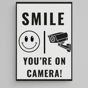 May include: Black and white framed sign with the text "SMILE" above a smiley face and a security camera icon. Below, the text reads "YOU'RE ON CAMERA!" The sign is rectangular and mounted on a light gray wall.