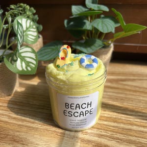 May include: A jar of yellow slime with a beach theme. The slime is topped with a miniature flip-flop and a blue and white life preserver. The label reads "BEACH ESCAPE" with scent and texture details.