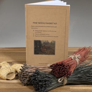 May include: A pine needle basket kit with a tan instructional booklet featuring the text PINE NEEDLE BASKET KIT, bundles of long pine needles in shades of red, brown, and dark green, and small coils of light tan cordage for stitching the basket.
